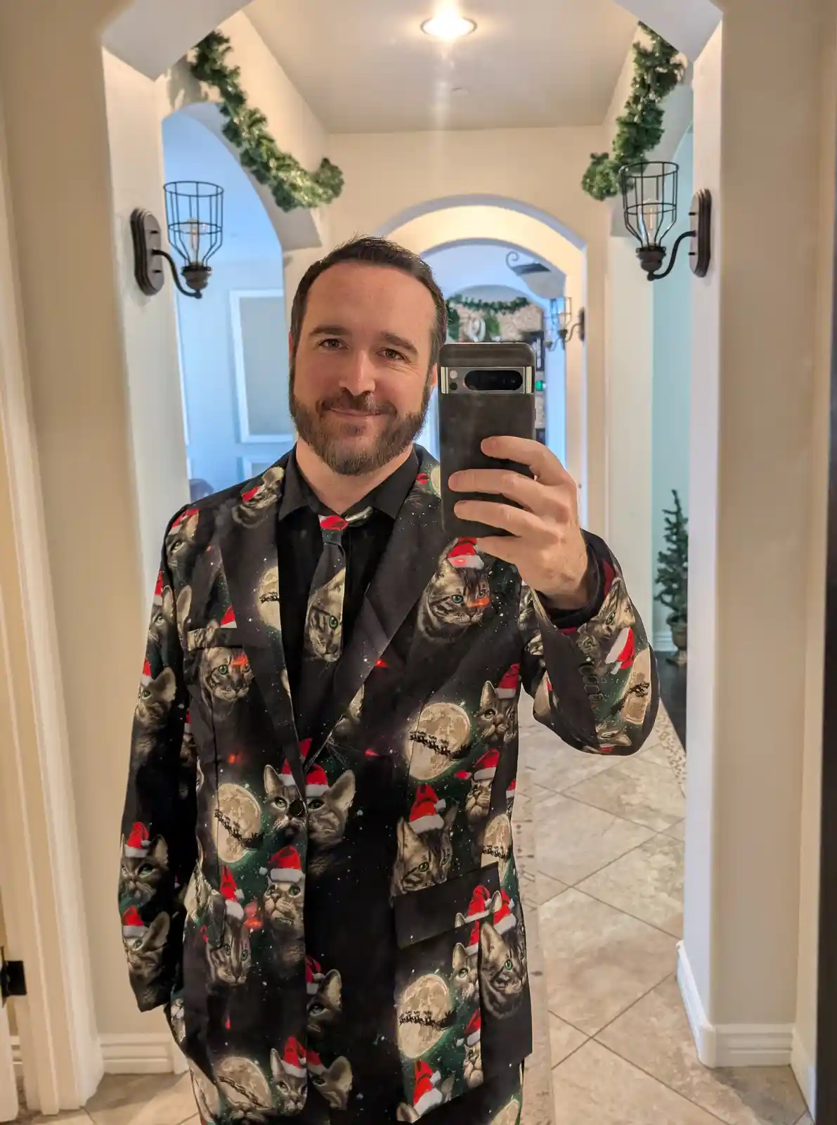 A man with a beard takes a mirror selfie in a hallway decorated with green garland. He wears a black blazer covered in a…