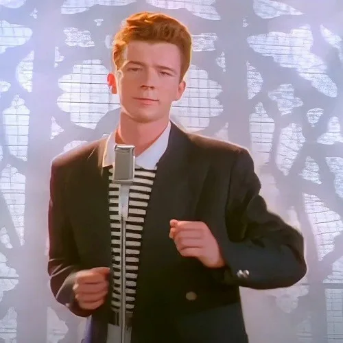 A man with red hair wearing a black blazer over a black-and-white striped shirt stands in front of a microphone, with a…