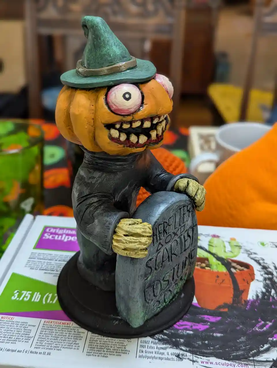 A handmade, orange pumpkin-headed figurine wearing a green witch hat and black robe, sitting on a tombstone that reads…