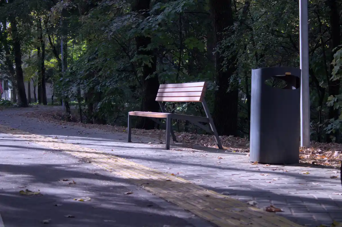 An empty wooden park bench sits beside a trash can on a paved path with yellow tactile paving, surrounded by trees with…