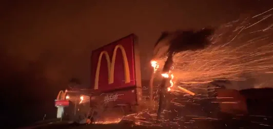 A nighttime image of a McDonald's restaurant engulfed in flames and sparks, with the iconic golden arches visible on the…