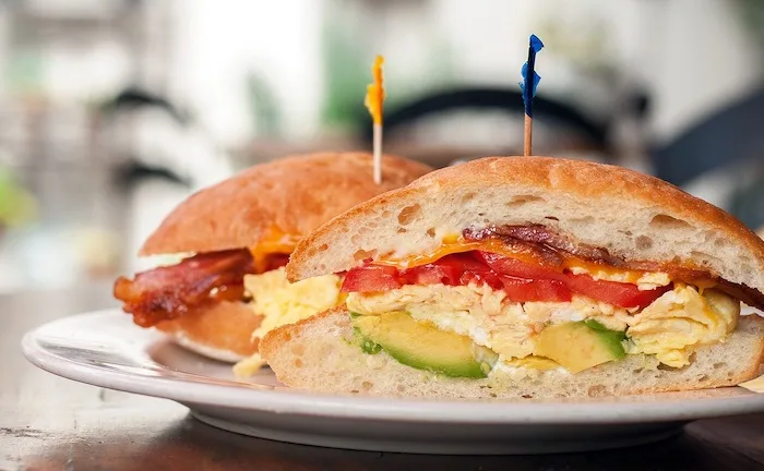 A close-up photo of two breakfast sandwiches on a plate, cut in half to show scrambled eggs, bacon, tomato, and avocado…
