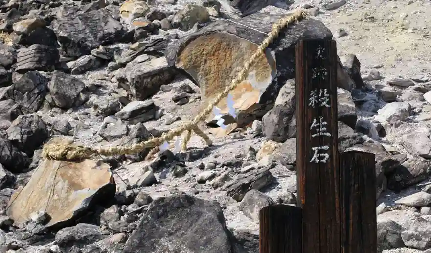 A wooden signpost with vertical Japanese characters '殺生石' stands in a rocky, barren landscape. A thick, braided rope…