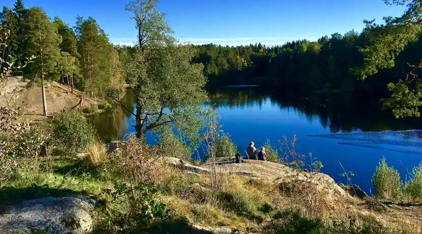 Two people sit on a rocky ledge overlooking a serene, deep blue lake surrounded by dense green forest under a clear blue…
