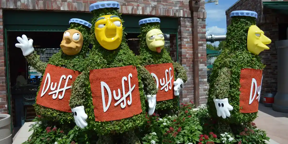 Four topiary sculptures shaped like Duff beer bottles, each with a Simpsons character's face (Homer, Moe, Barney, and a…
