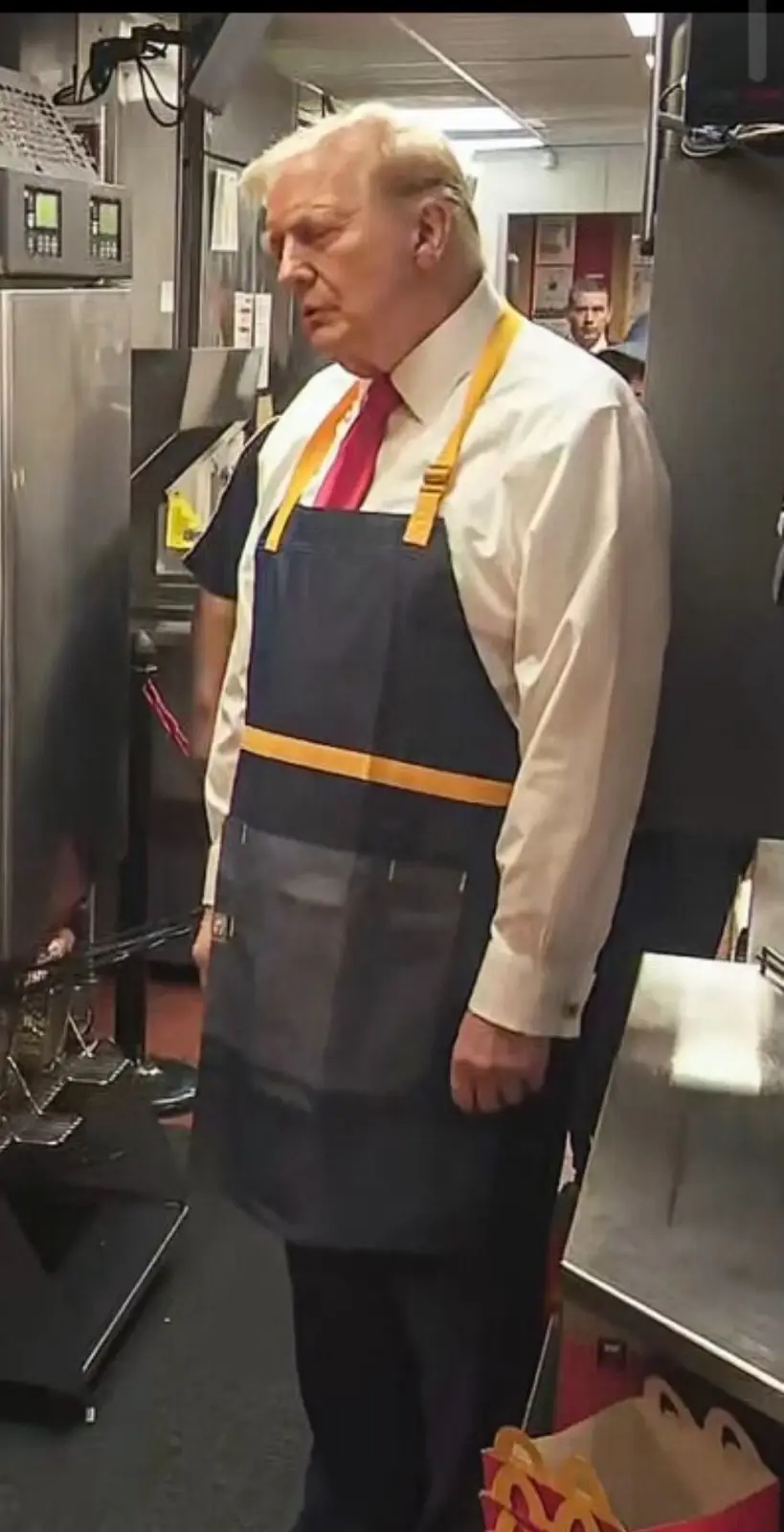 A man with white hair and a red tie wears a dark blue McDonald's apron with yellow straps in a kitchen setting. He stands…
