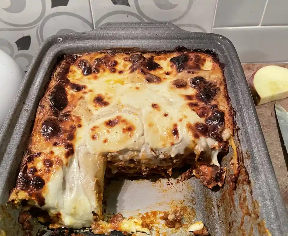 A close-up photo shows a grey baking dish containing a lasagna with a golden-brown, slightly burnt cheese topping. A square…