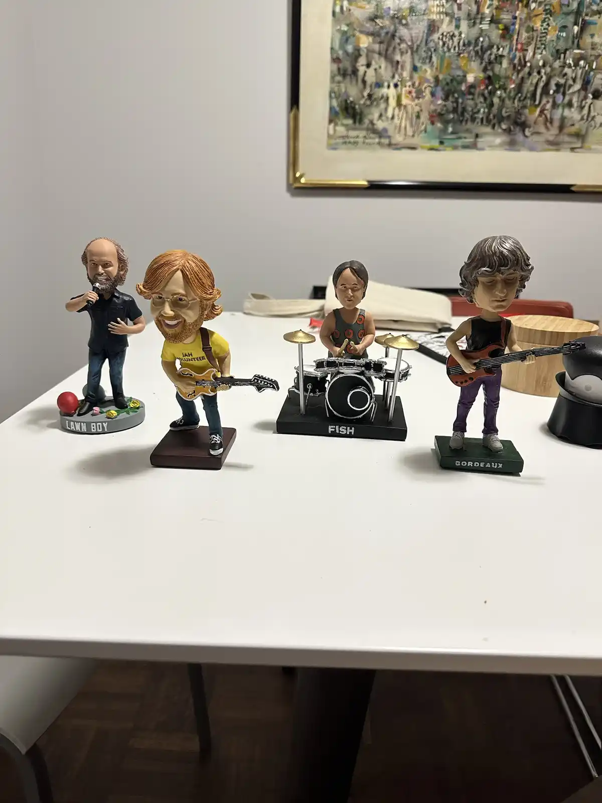 Four bobblehead figurines of musicians are arranged on a white table, each holding an instrument: a singer with a mic, a…