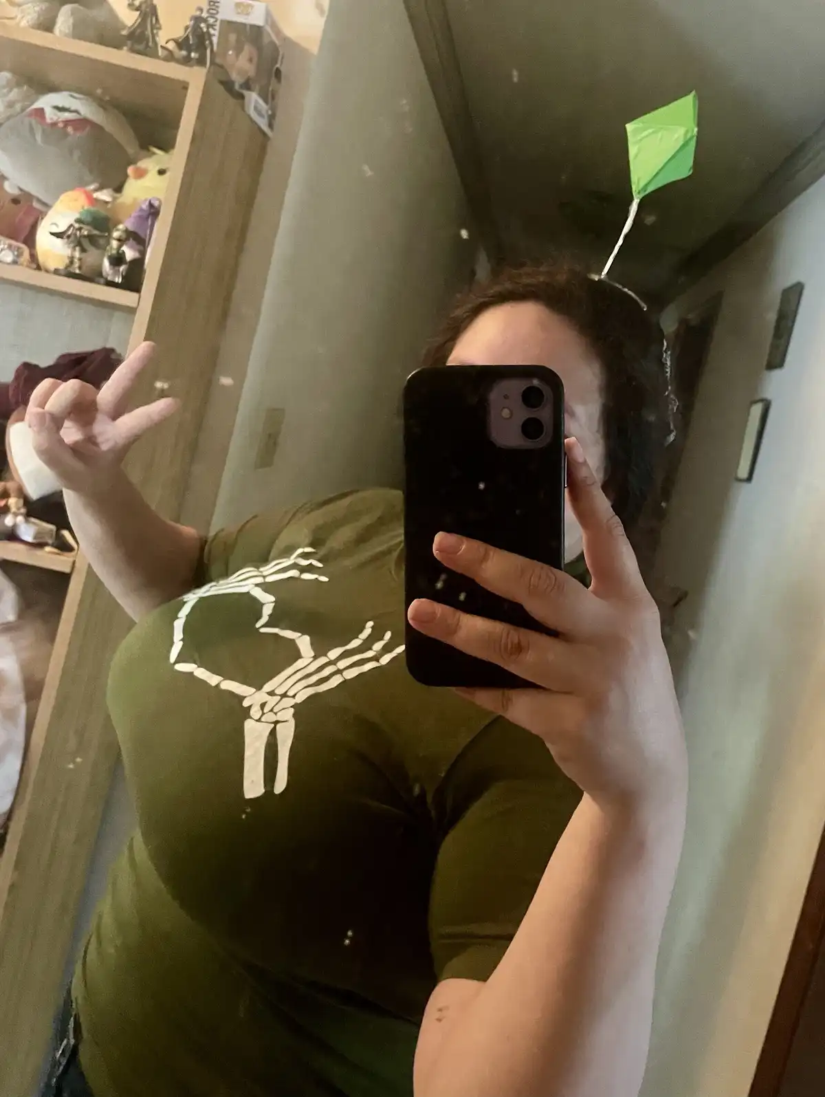 A person takes a mirror selfie holding a black smartphone, wearing a green t-shirt with a white skeleton hand graphic, and…