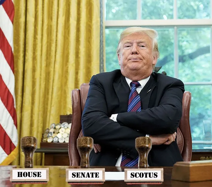 A man in a dark suit and striped tie sits with arms crossed at a desk in an office with yellow curtains and an American…
