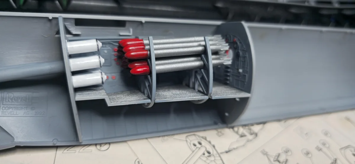 A close-up of a grey plastic model kit's internal compartment, showing three white cylindrical parts, a cluster of…