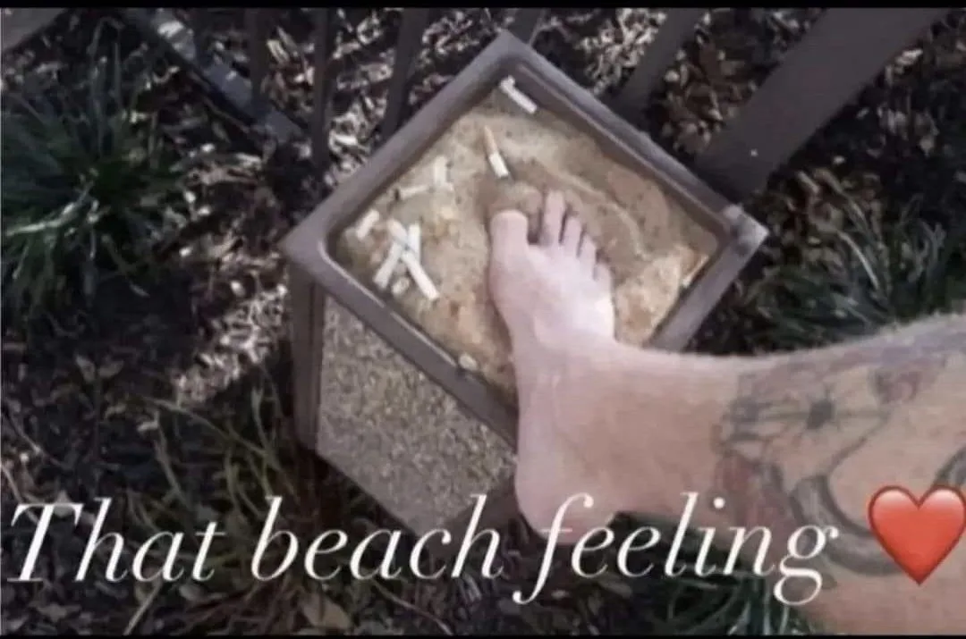 A person's bare foot is pressed into a square sand-filled planter, surrounded by cigarette butts. The person has a tattooed…