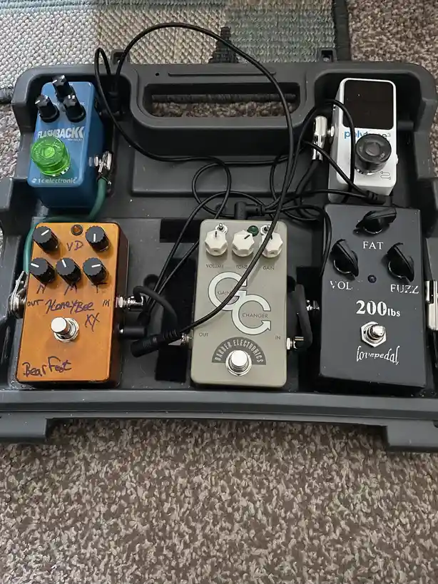 A top-down photo of four guitar effect pedals arranged in a black pedalboard case on carpet. The pedals include a blue TC…