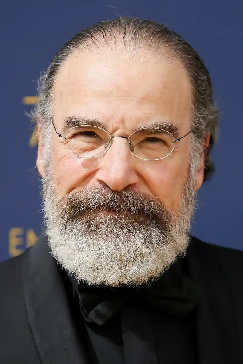 A close-up photo of a man with a gray beard and round glasses, wearing a black tuxedo and bowtie, looking directly at the…