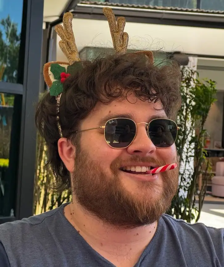 a man with curly brown hair and a beard wears gold-rimmed sunglasses and a glittery reindeer antler headband with a holly…