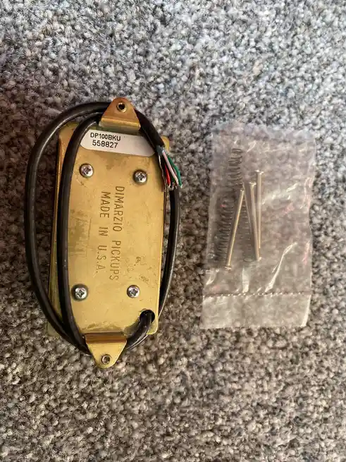 A brass-colored Dimarzio guitar pickup with black wiring and a white sticker reading 'DP100BKF 558827' lies on a gray carpet…