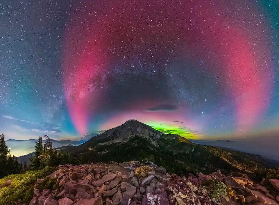 A stunning night-time photograph shows a mountain range under a starry sky with vibrant pink and green auroras. The…