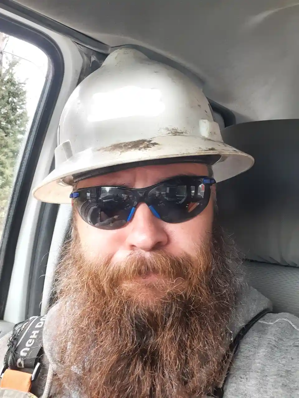 A man with a long brown beard wearing a dirty white hard hat and dark sunglasses sits inside a vehicle, looking directly at…