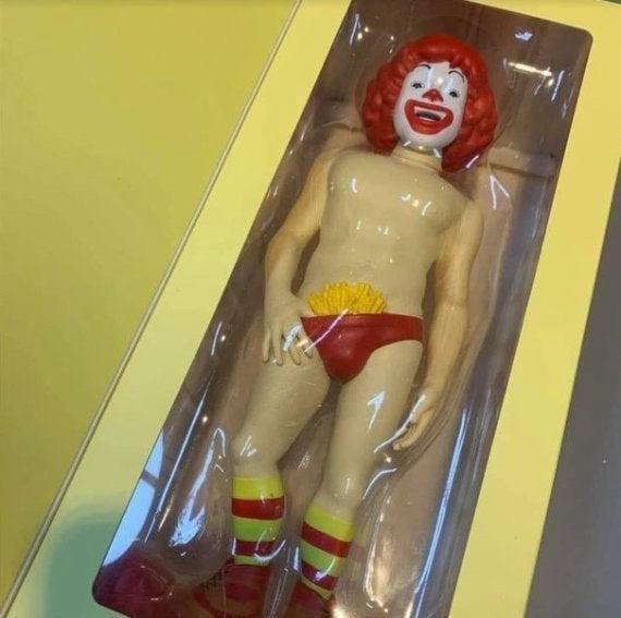 A Ronald McDonald action figure is shown in its original clear plastic packaging, standing upright. The figure is unclothed…