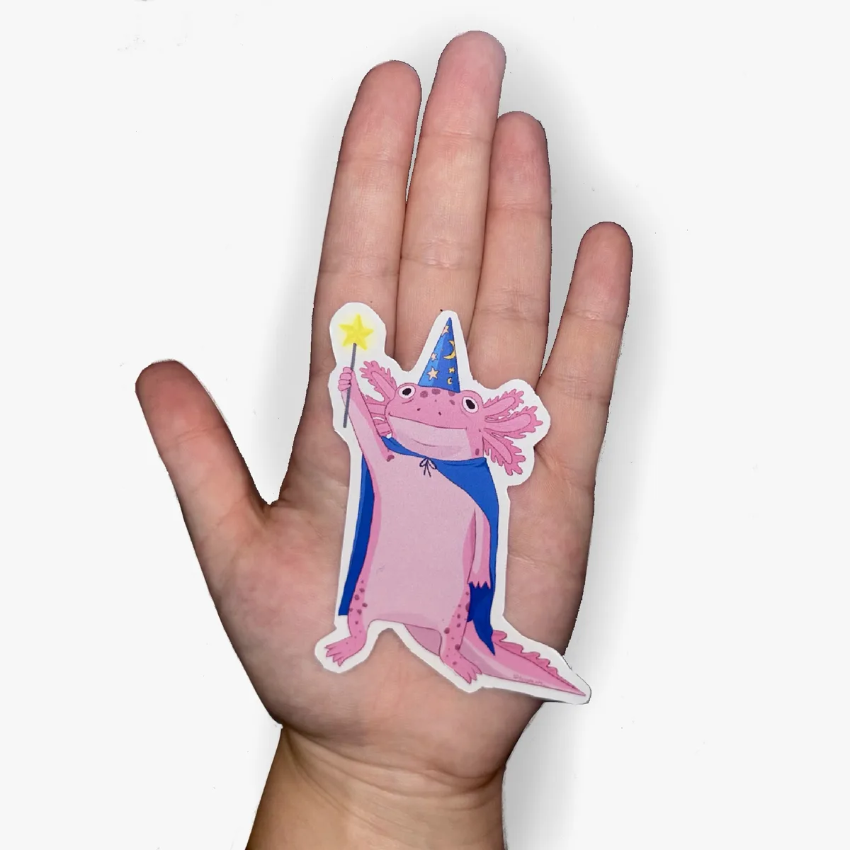 A hand holds a pink axolotl sticker dressed as a wizard, wearing a blue pointed hat with stars and moons, a blue cape, and…