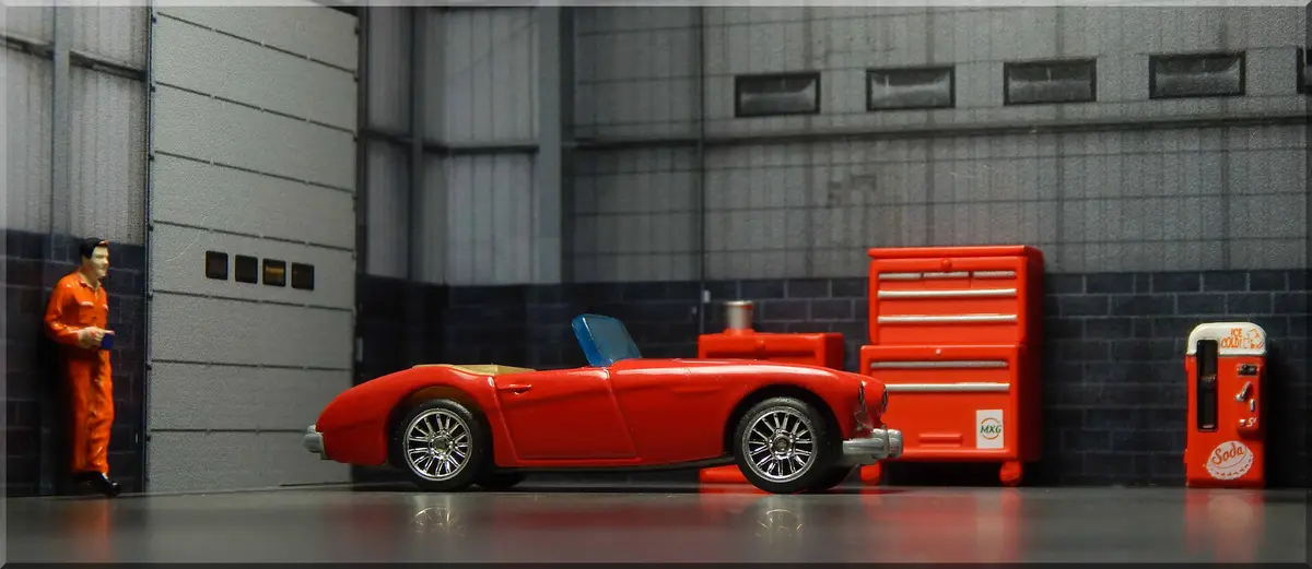 A miniature scene depicts a red toy sports car parked in a garage next to a red toolbox and a soda vending machine. A small…