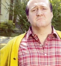a man with a grumpy, unimpressed facial expression wears a red plaid shirt under a yellow cardigan, standing outdoors with…