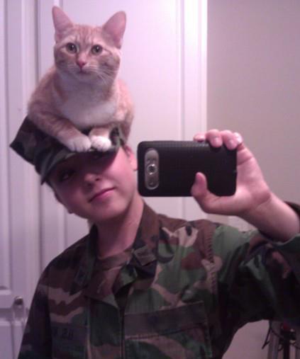 A young person in a camouflage military-style uniform and cap is taking a selfie with a phone, while a ginger and white cat…