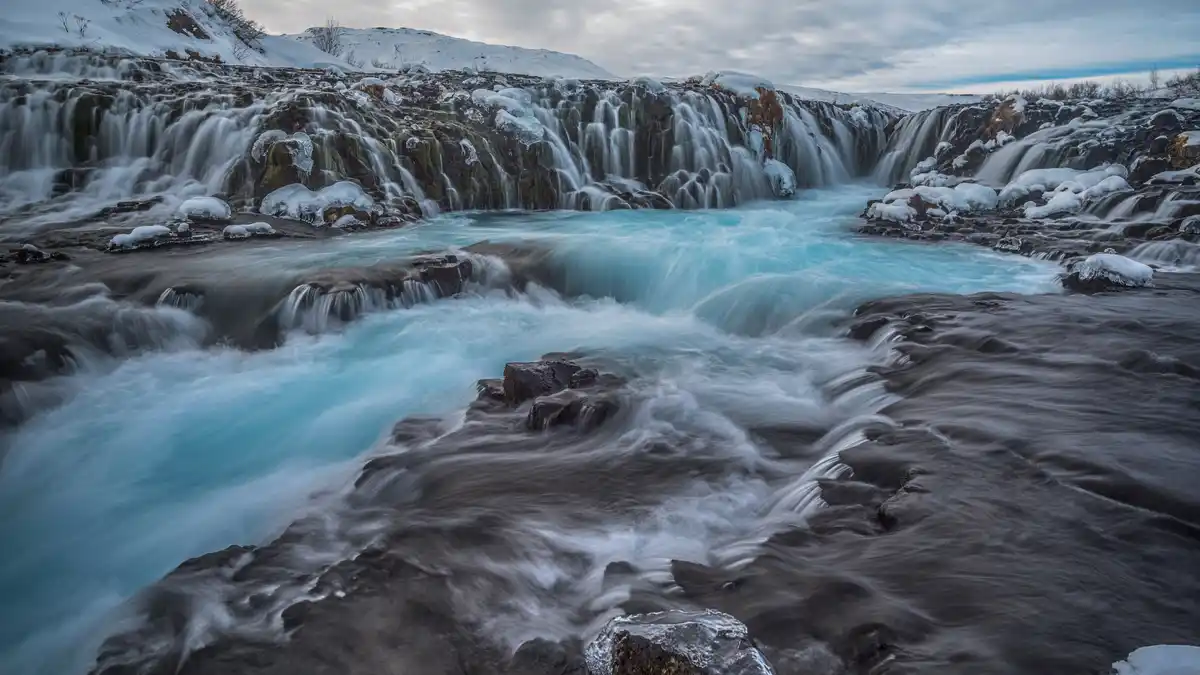 A long-exposure photograph captures a wide, icy waterfall cascading over dark, snow-dusted rocks under an overcast sky, with…