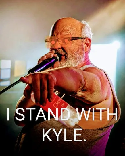 A bald, bearded man with glasses, wearing a tank top, sings into a microphone while holding a red bottle. The text overlaid…