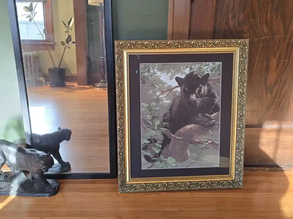 A framed painting of a black panther resting in a tree is propped up on a wooden floor next to a black panther statue. The…