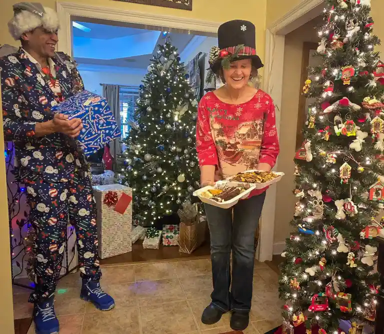 Two people in a festive, decorated home stand near Christmas trees. The man on the left wears a full-body holiday-themed…