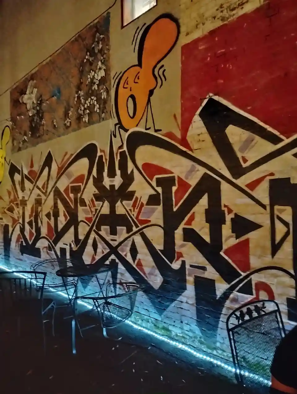 A photo of a brick wall covered in colorful graffiti, featuring a large orange cartoon character with an exaggerated yelling…
