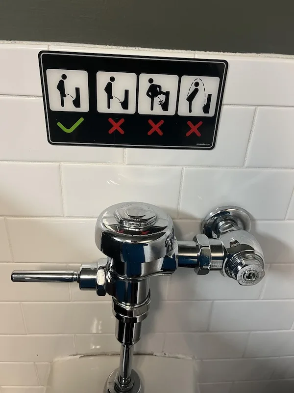 A close-up photo of a chrome urinal flush valve mounted on a white tiled wall, with a black instructional sign above it. The…
