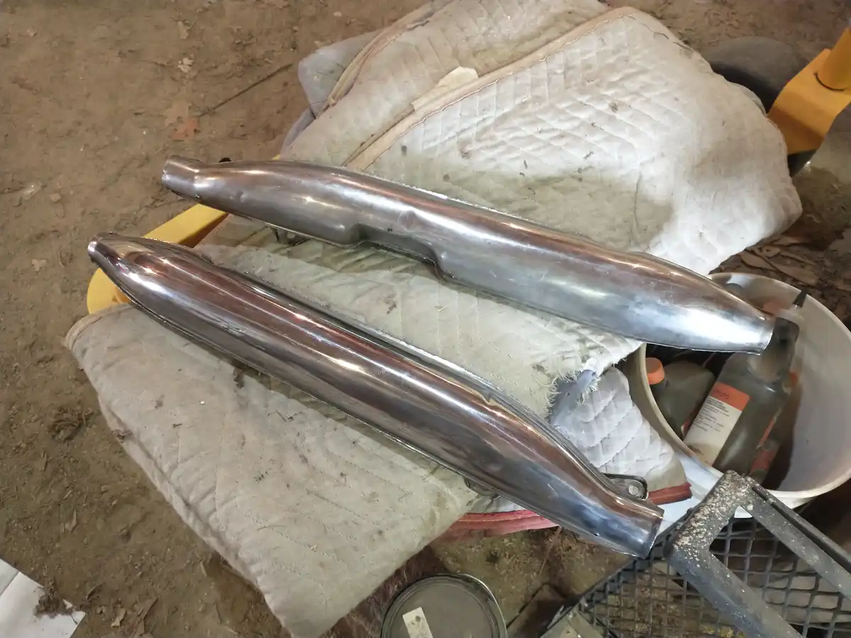 Two shiny, polished metal exhaust pipes rest on a dirty quilted mat in a cluttered garage, with tools and containers nearby…