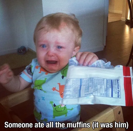 A baby with blonde hair and a blue shirt with green monster prints is visibly crying while holding a partially eaten muffin…