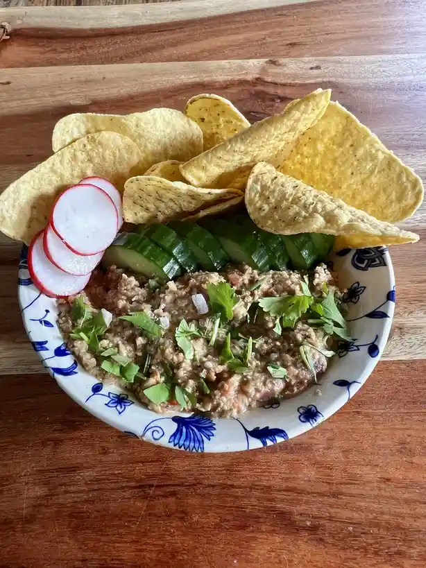 A bowl of textured dip, garnished with cilantro, sits on a wooden surface with tortilla chips, sliced radishes, and cucumber…