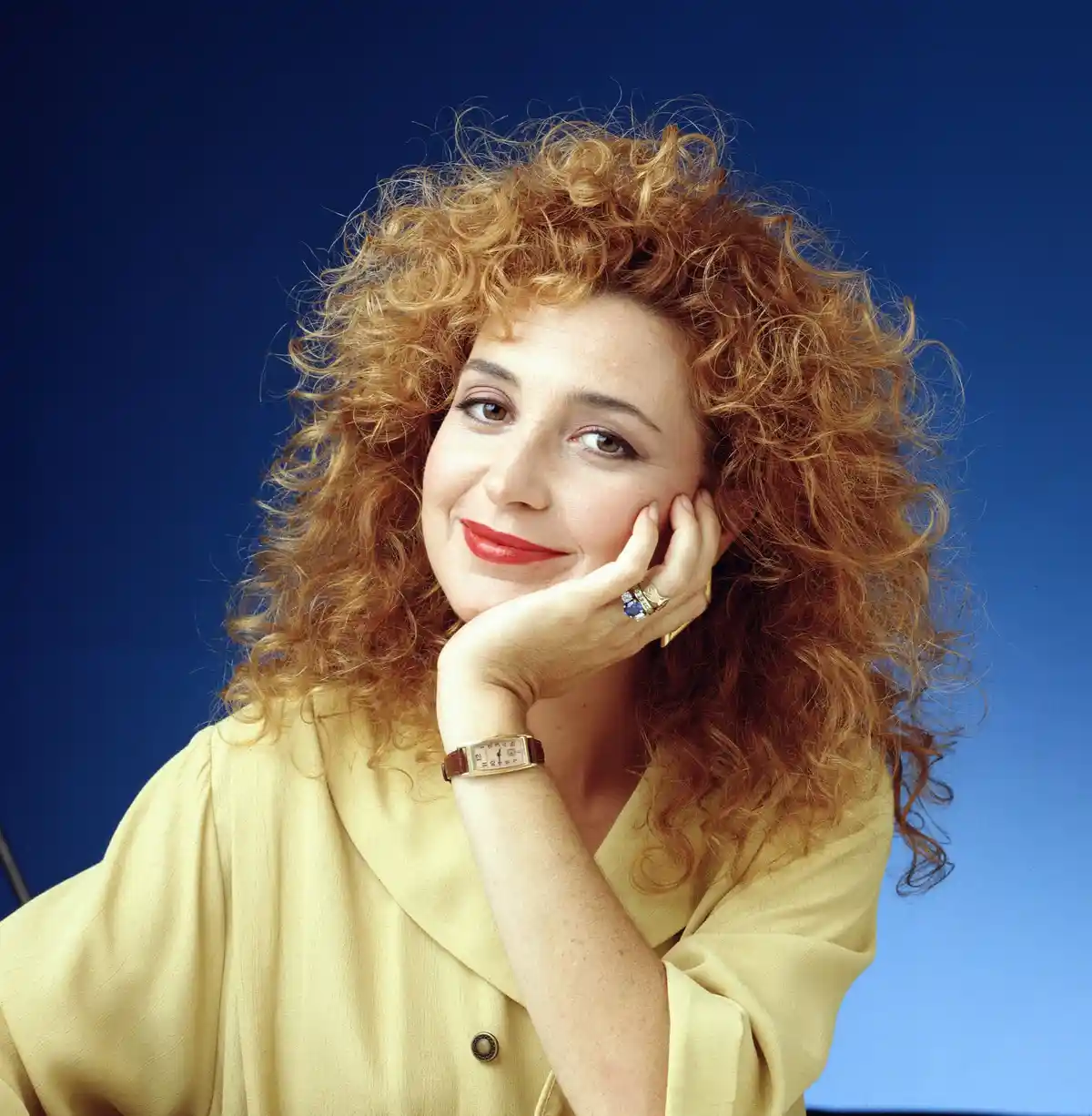 a woman with voluminous curly red hair and red lipstick poses against a solid blue background, resting her chin on her hand…