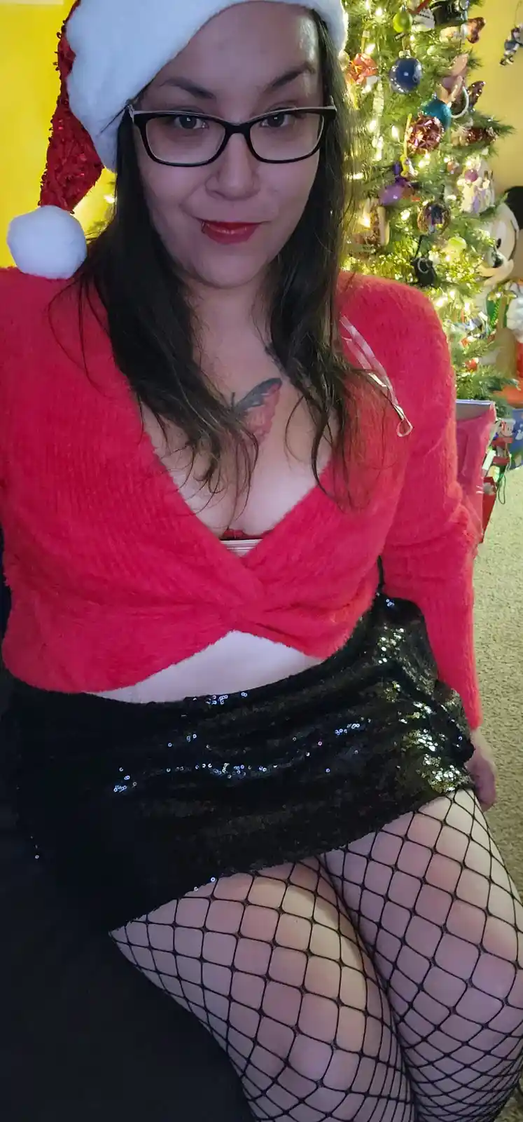 A woman wearing a red fuzzy Santa-style top, black sequined skirt, fishnet stockings, and a Santa hat poses in front of a…