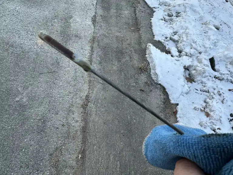 A person wearing a blue work glove holds a long metal dipstick against a concrete sidewalk next to melting snow. The…