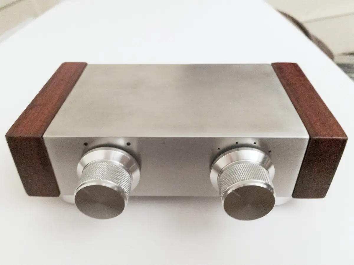 A close-up photo of a minimalist audio device with a brushed metal body and dark wood side panels, featuring two large…