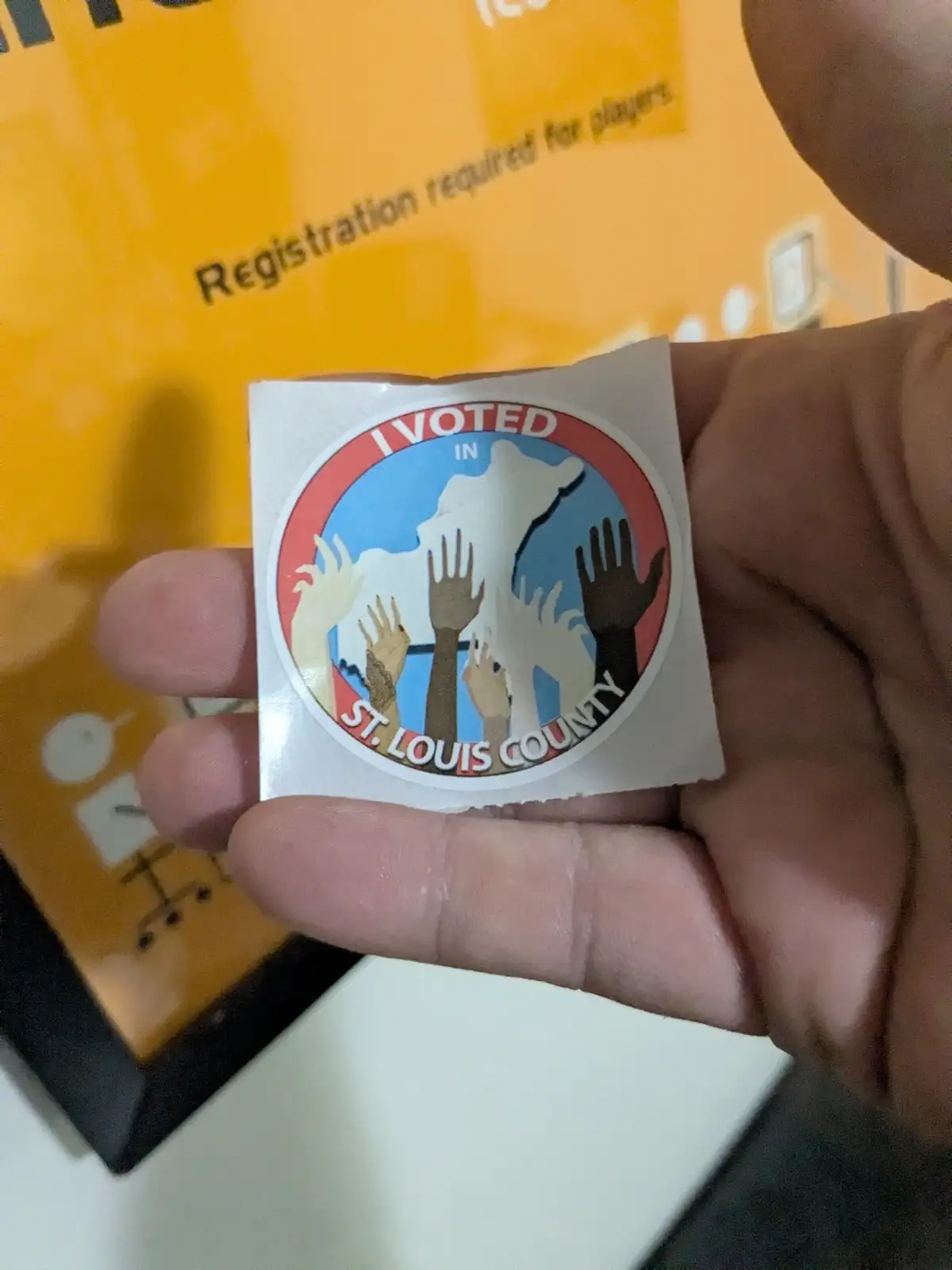 A hand holds a circular 'I VOTED IN ST. LOUIS COUNTY' sticker featuring diverse hands reaching upward against a blue…