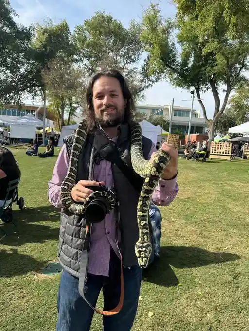 A man with long dark hair and a beard, wearing a purple checkered shirt and grey vest, stands outdoors in a grassy park…