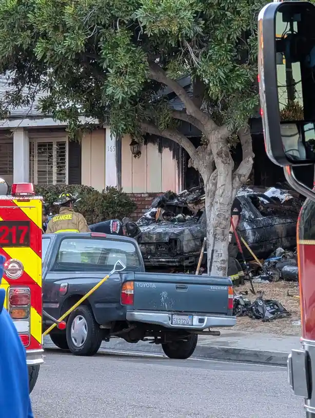 A photo shows firefighters at a house fire scene with a severely burned car behind a tree. A dark blue Toyota Tacoma pickup…