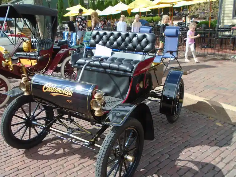 A vintage 1904 Oldsmobile car with black and red paint, brass headlights, and tufted leather seats is parked on a brick…