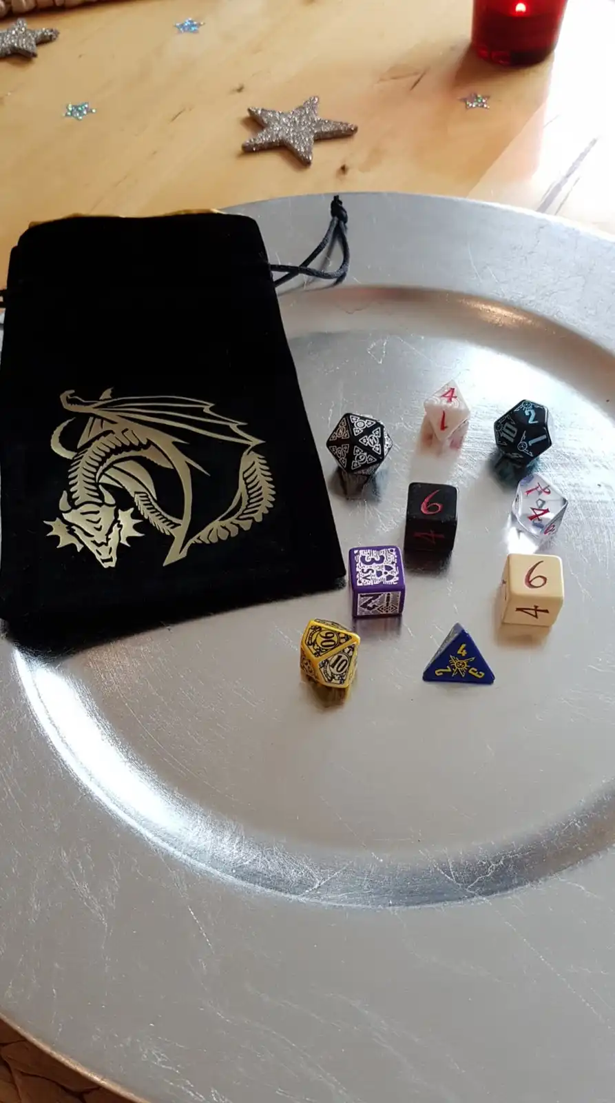 A silver tray holds a collection of colorful polyhedral dice, mostly showing 4s and 6s, next to a black velvet bag with a…