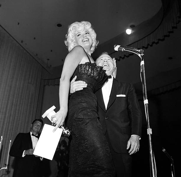 A black-and-white photo shows Marilyn Monroe laughing while being held up by a man in a tuxedo on a stage with a microphone…