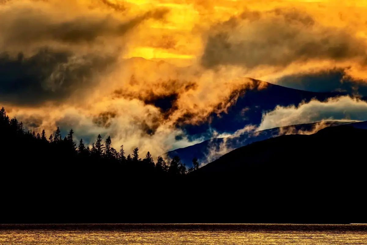 A dramatic landscape photo shows a golden sunset or sunrise with fiery orange and yellow clouds illuminating mountain peaks…