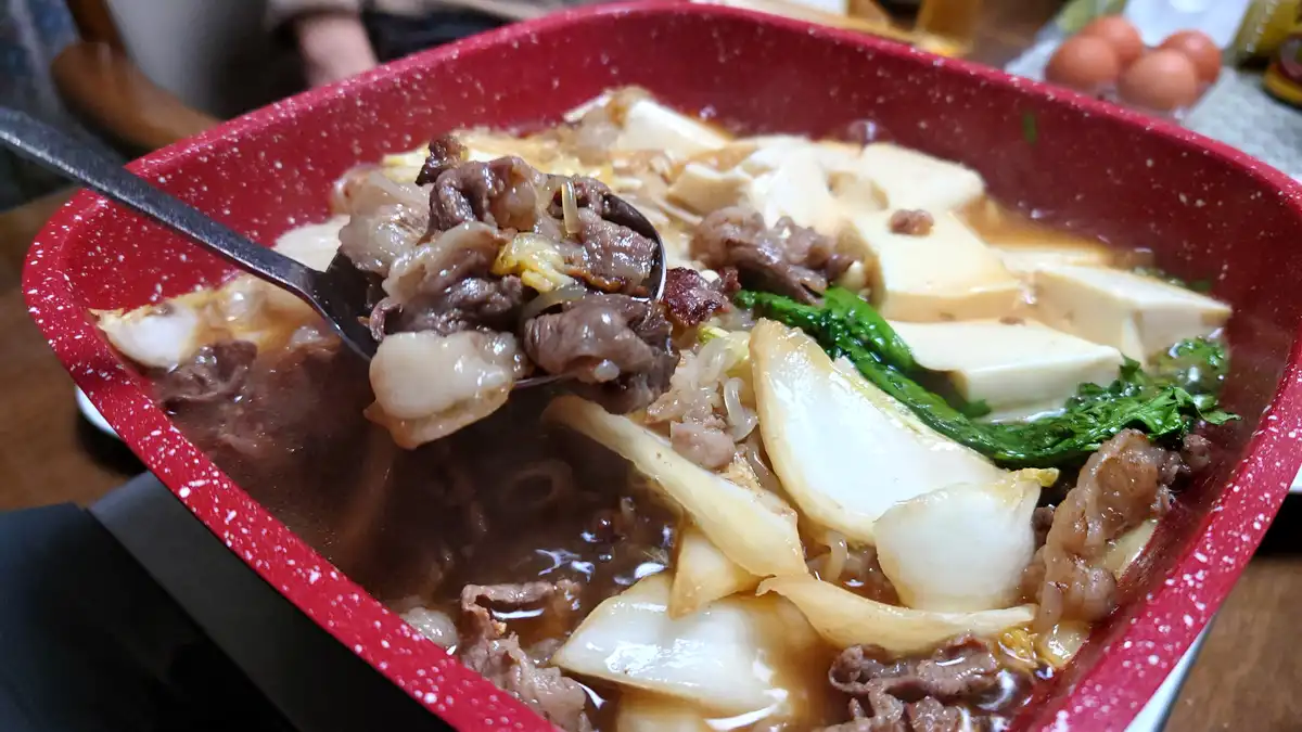 a close-up photo of a red speckled rectangular pot filled with beef hot pot, showing chunks of beef, tofu, cabbage, and…