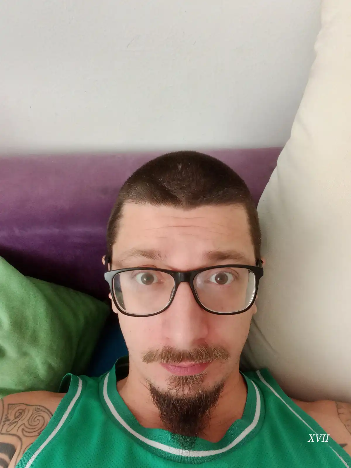A man with short dark hair, glasses, and a goatee wearing a green tank top lies on a bed with purple and green pillows…