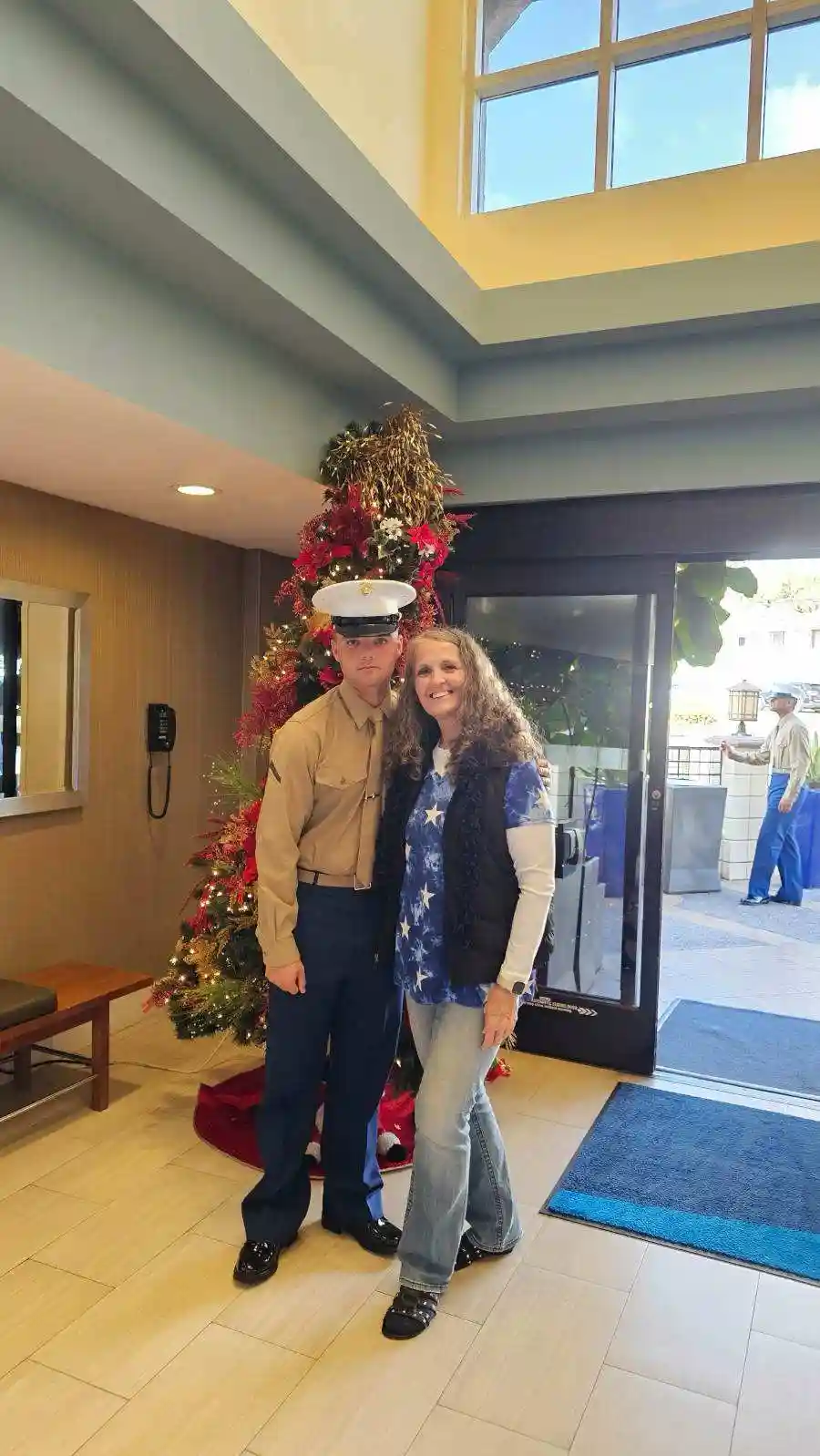 A man in a U.S. Marine Corps dress uniform stands stiffly beside a smiling woman in casual clothes, both posing for a photo…
